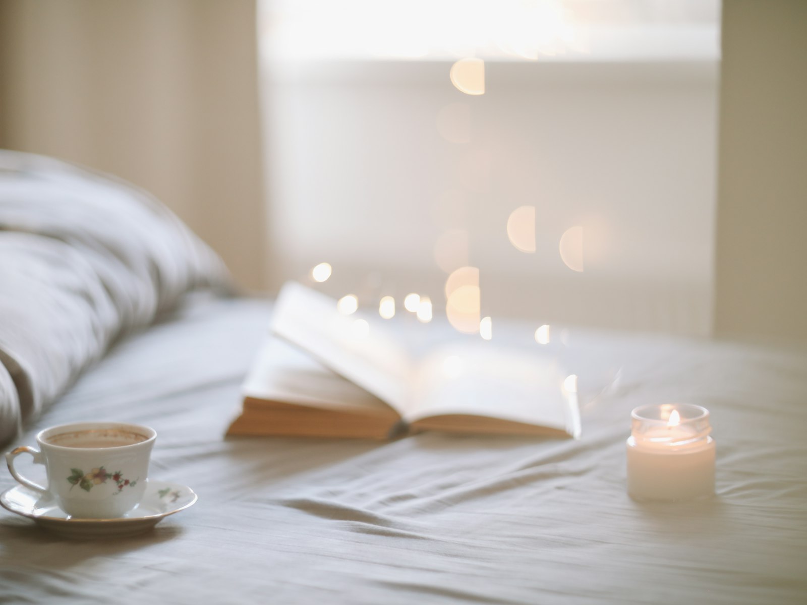 Open book, tea cup and a candle on a bed with warm lights behind