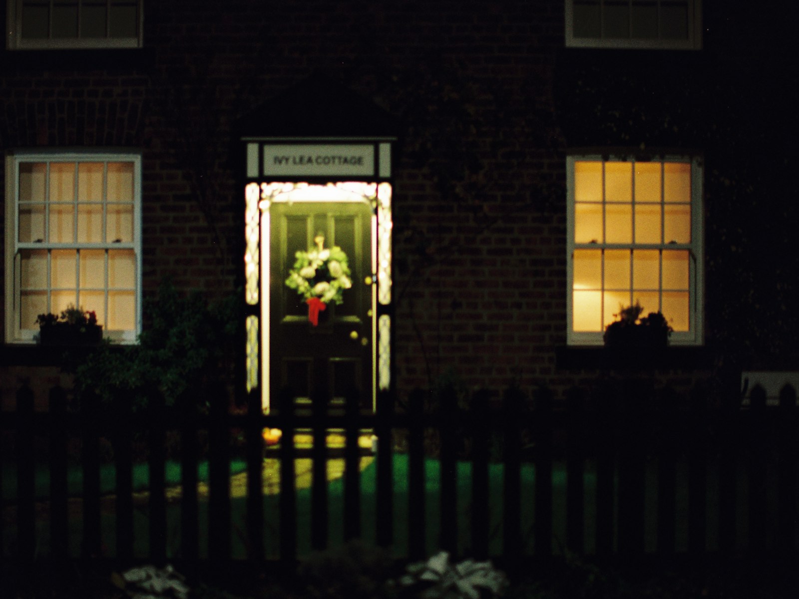 Front door with a lit wreath glowing at dusk between lit windows