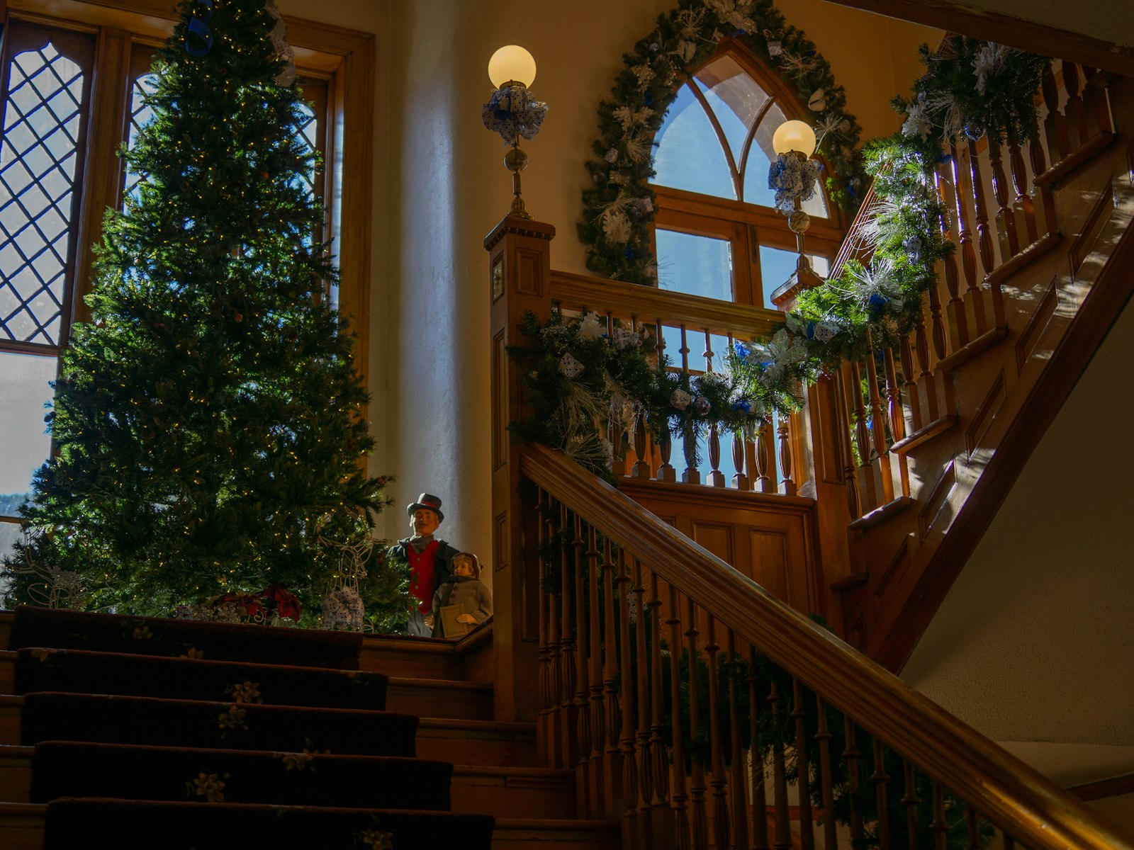 Wood staircase with garland beside a lit Christmas tree in an entry hall