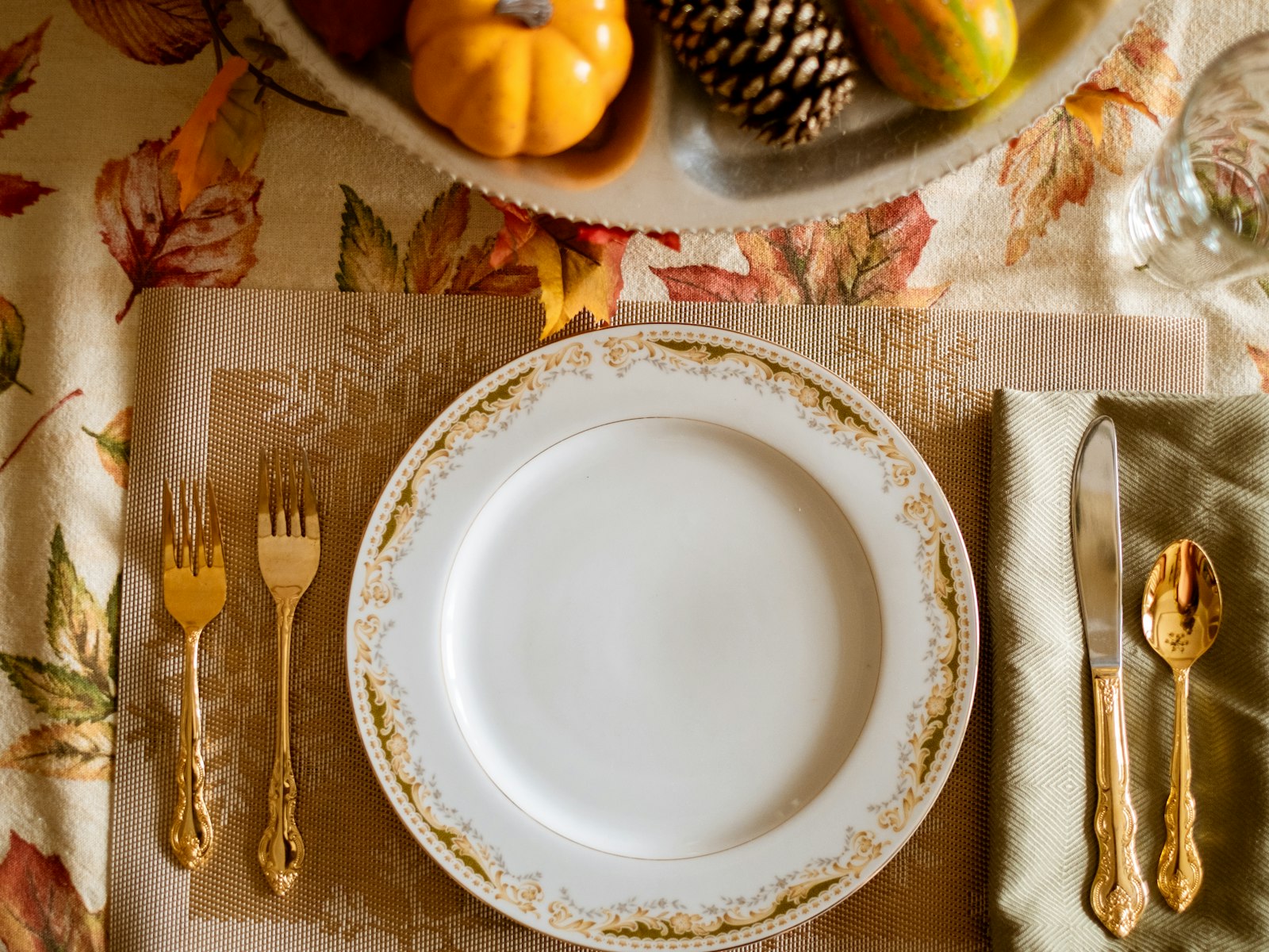 Thanksgiving place setting with a gold-rimmed plate, gold flatware and a linen napkin on an autumn leaf tablecloth